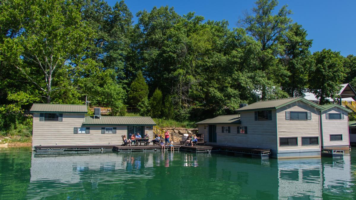 Floating cabins on a calm lake surrounded by lush green trees, with people relaxing on the docks