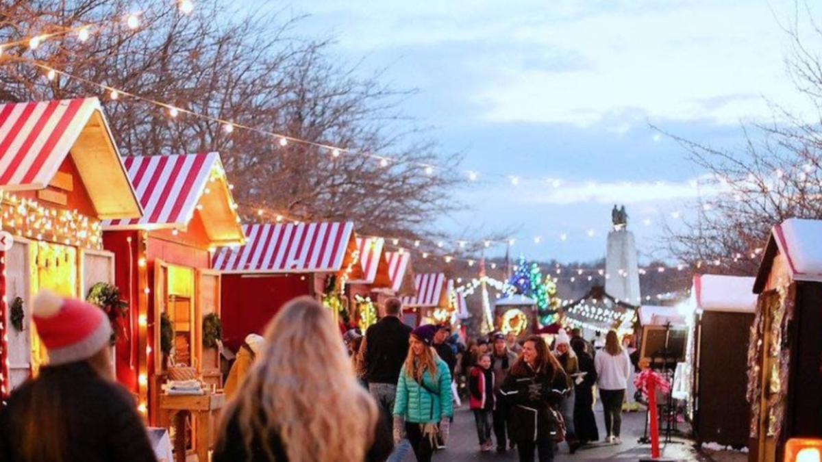 A festive outdoor Christmas market at dusk, featuring small wooden booths with red and white striped roofs, adorned with string lights. Visitors stroll through the lively market, bundled in warm coats and hats, enjoying the holiday ambiance under a sky turning from blue to twilight. In the background, more lights and a tall monument or statue can be seen, adding to the magical winter atmosphere.
