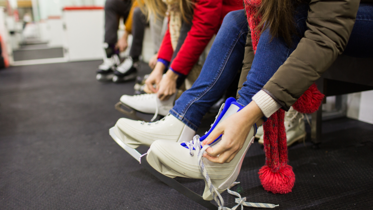 A close-up view of people sitting on a bench, lacing up their ice skates in preparation for skating. The person in the foreground wears a brown winter coat, blue jeans, and a red scarf, tying the laces of white ice skates. Others in the background are also getting ready, creating a lively, anticipatory scene at an indoor or outdoor skating rink.