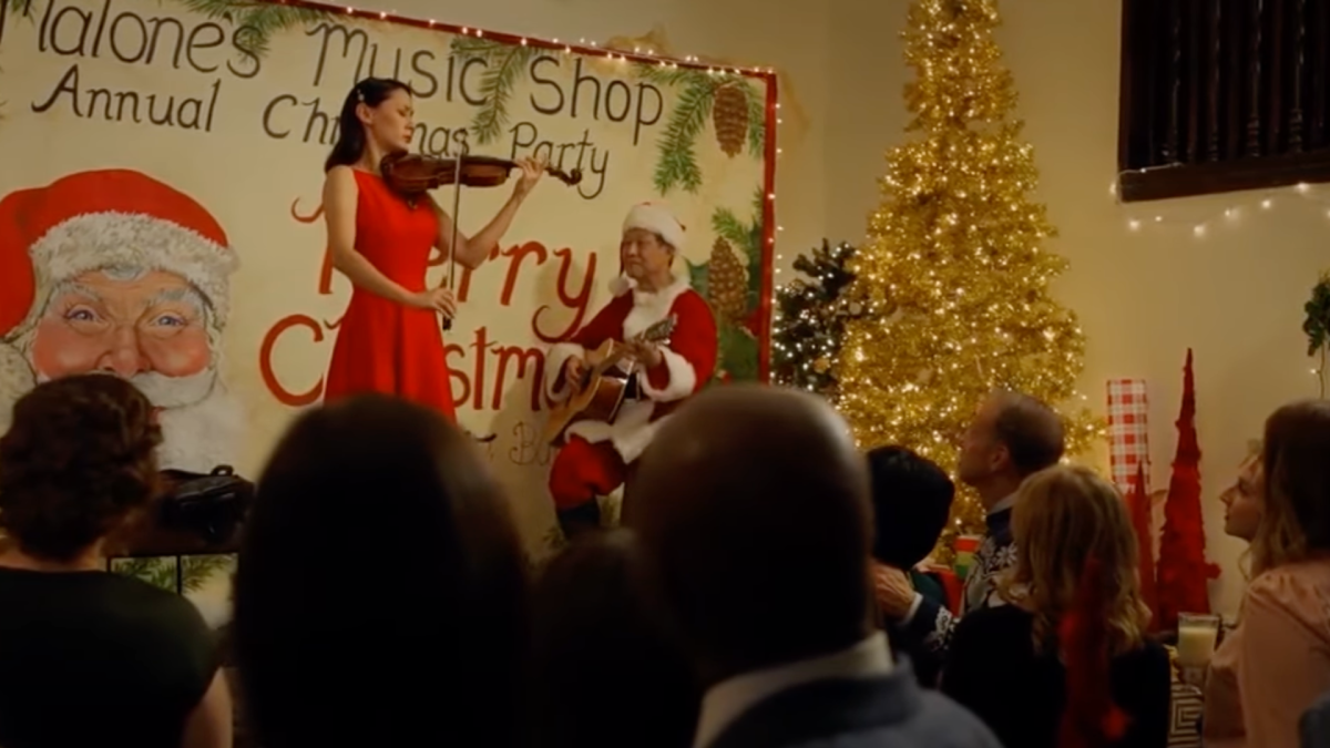 A festive indoor Christmas concert scene featuring a woman in a red dress playing the violin, standing in front of a decorated backdrop that reads "Malone's Music Shop Annual Christmas Party." A man dressed as Santa Claus accompanies her on guitar. The stage is adorned with holiday decor, including a large glowing Christmas tree and festive lights, while a joyful audience watches and listens attentively.