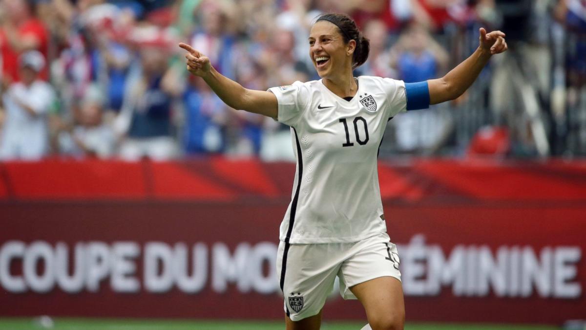 Carli Lloyd wears number 10 on her USWNT white home uniform, she is smiling with her arms stretched wide and points to the crowd