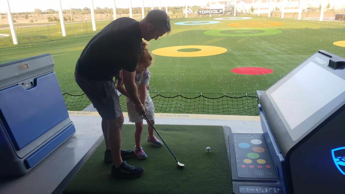 A father leans over to help his daughter hit a golf ball at Topgolf Wichita