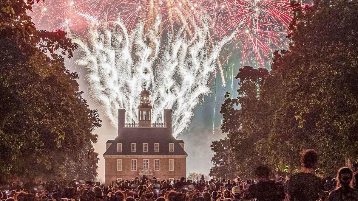 Fireworks illuminate the night sky above a historic building, surrounded by a crowd of spectators. Lush trees frame the scene, enhancing the festive atmosphere.