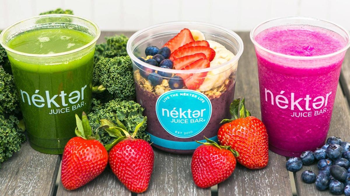The camera faces a wooden picnic table featuring three plastic cups bearing the "nékter JUICE BAR" label. The cup on the left is filled with a bright green smoothie and surrounded by broccoli stalks. The cup in the middle contains a dark purple-ish smoothie topped with oats, blueberries, strawberries, and banana slices. The cup on the right is full of a bright magenta (dragonfruit?) smoothie surrounded by strawberries and blueberries.