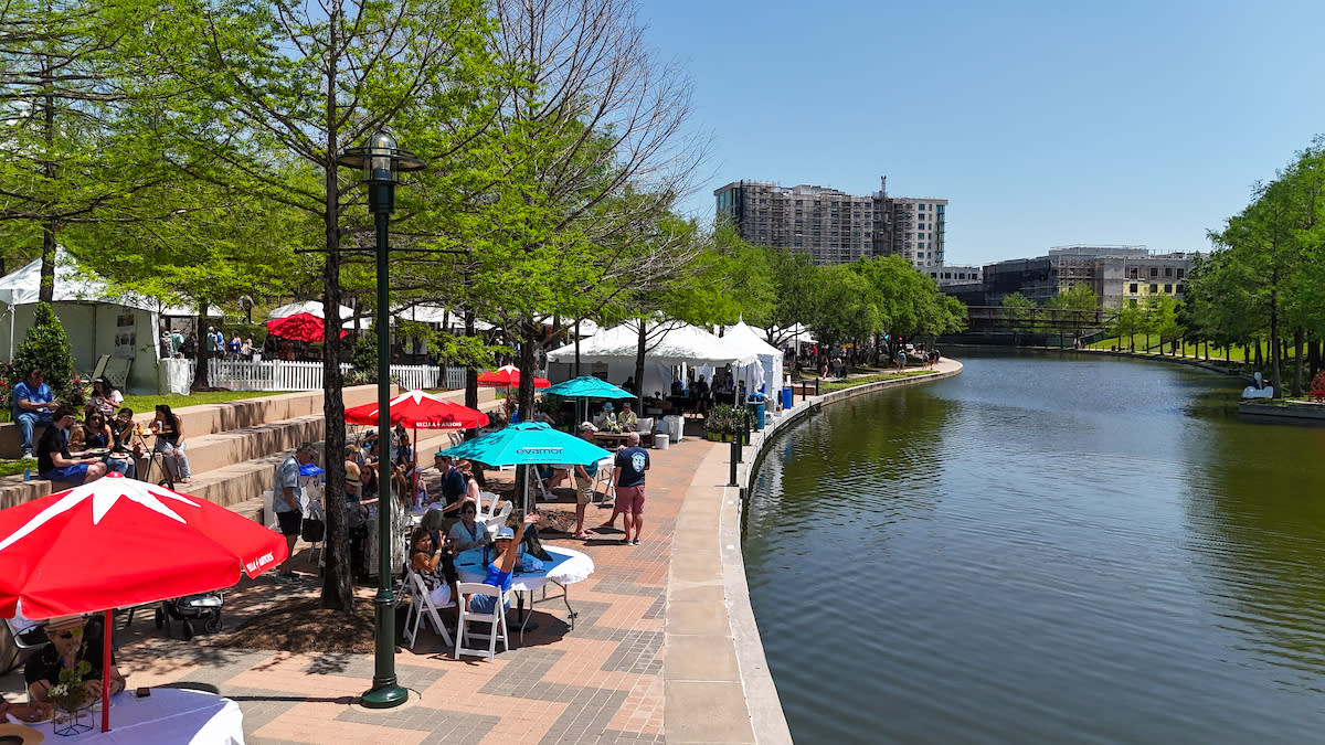The camera focuses on the VIP area of the WWAF, featuring several tables shaded by colorful umbrellas or white pop-up tents. This area is right along the Waterway. The day is bright and sunny without a cloud in the sky. A few office buildings are visible in the distance.