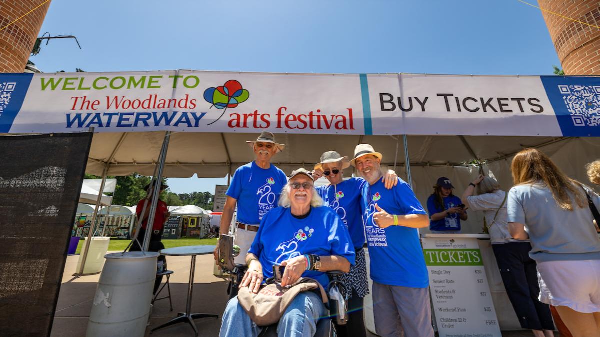 The entrance of Town Green Park and The Woodlands Waterway Arts Festival 2025 features two red-brick pillars topped with large metal pinecones. Four male volunteers smiling at the camera in front of the ticket booth.