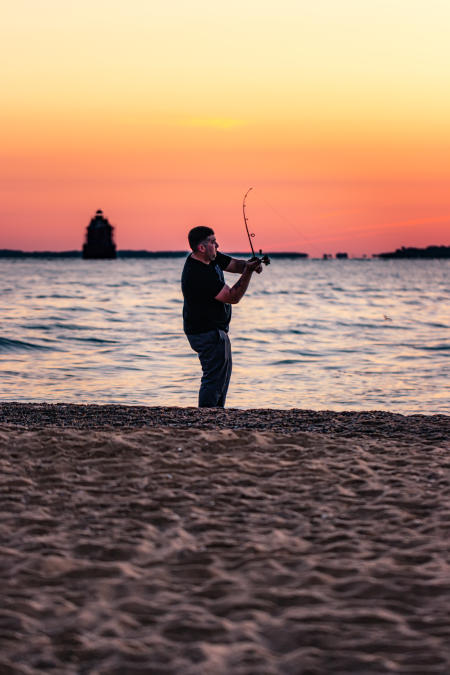 A man at a beach fishes from the shore with the sun setting in th ebackground.