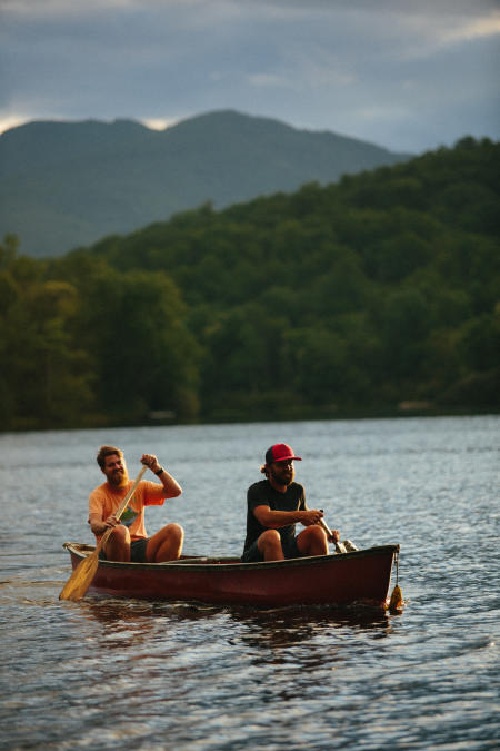 Two men sit in a canoe paddling on a lake. There are rolling green mountains behind the lake in the background.