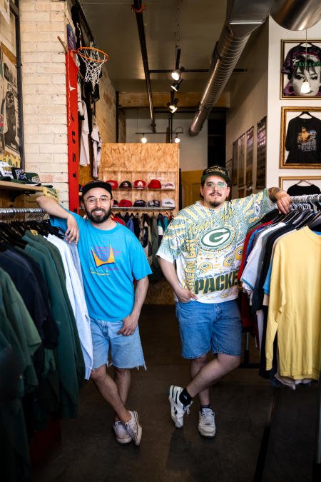 Two men standing in an isle at Manana Vintage in Green Bay, Wisconsin looking at the camera and resting their arms on clothing racks full of vintage t-shirts and sweatshirts.