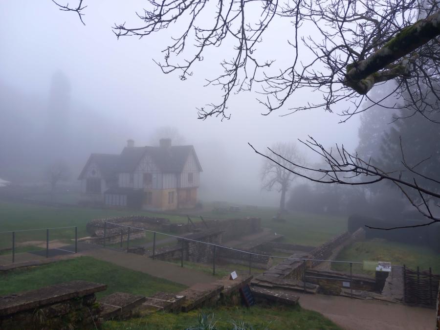 Chedworth Roman Villa in the Cotswolds on a misty morning, with historic buildings and archaeological remains partly obscured by fog.