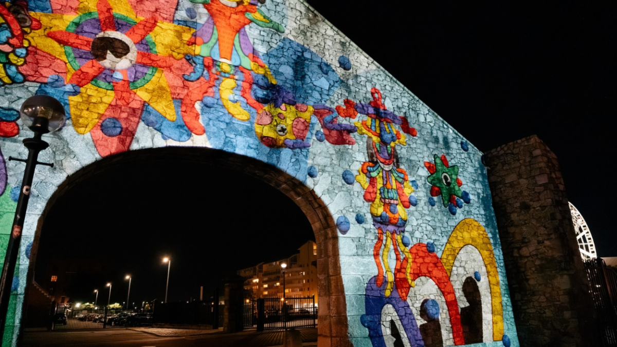 A colourful projection artwork covers a large stone archway at night, transforming the surface with vibrant patterns, abstract shapes and playful underwater-inspired illustrations in shades of blue, red, yellow, green and orange. The illuminated design contrasts against the dark sky, while nearby buildings and streetlights frame the waterfront setting.