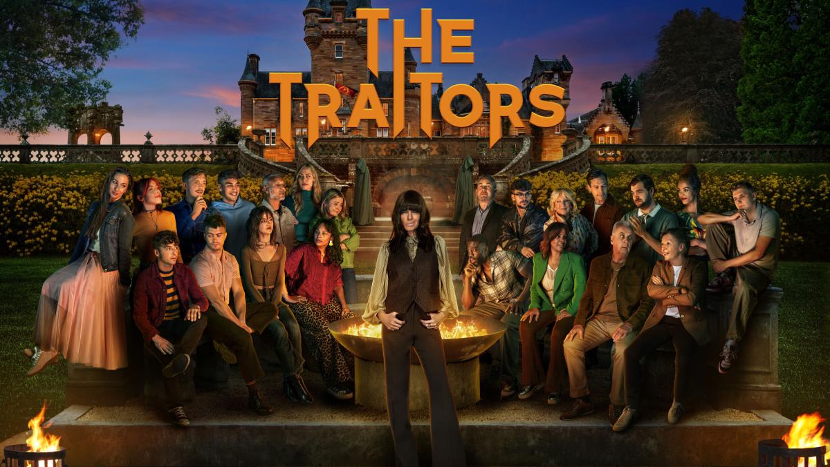 Promotional image for The Traitors featuring a large group of contestants gathered outdoors around a fire pit at night. A person stands in the centre foreground, with other participants seated and standing around them in a landscaped courtyard. Historic buildings and illuminated windows form the backdrop, while the programme title “The Traitors” appears in large orange lettering at the top of the image.