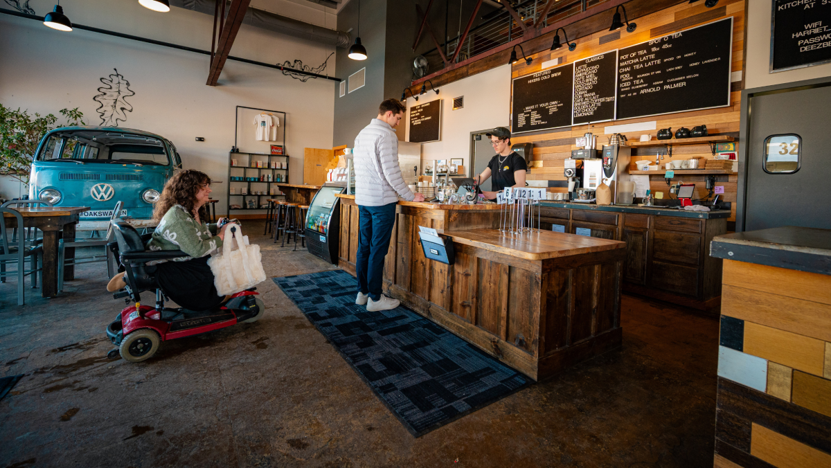 A cozy café with a barista behind a rustic wooden counter, a person in a white jacket ordering, and a customer in a wheelchair exploring the space. Modern and inviting.