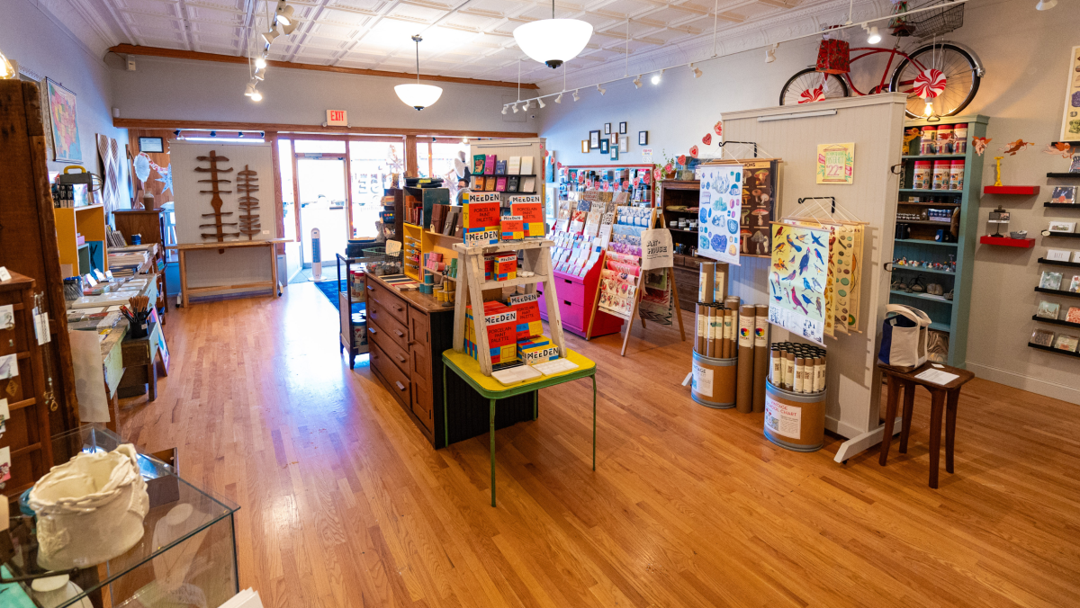 Cozy, well-lit store with wooden floors, displaying colorful stationery, art supplies, and greeting cards on neatly arranged shelves and tables. Welcoming ambiance.