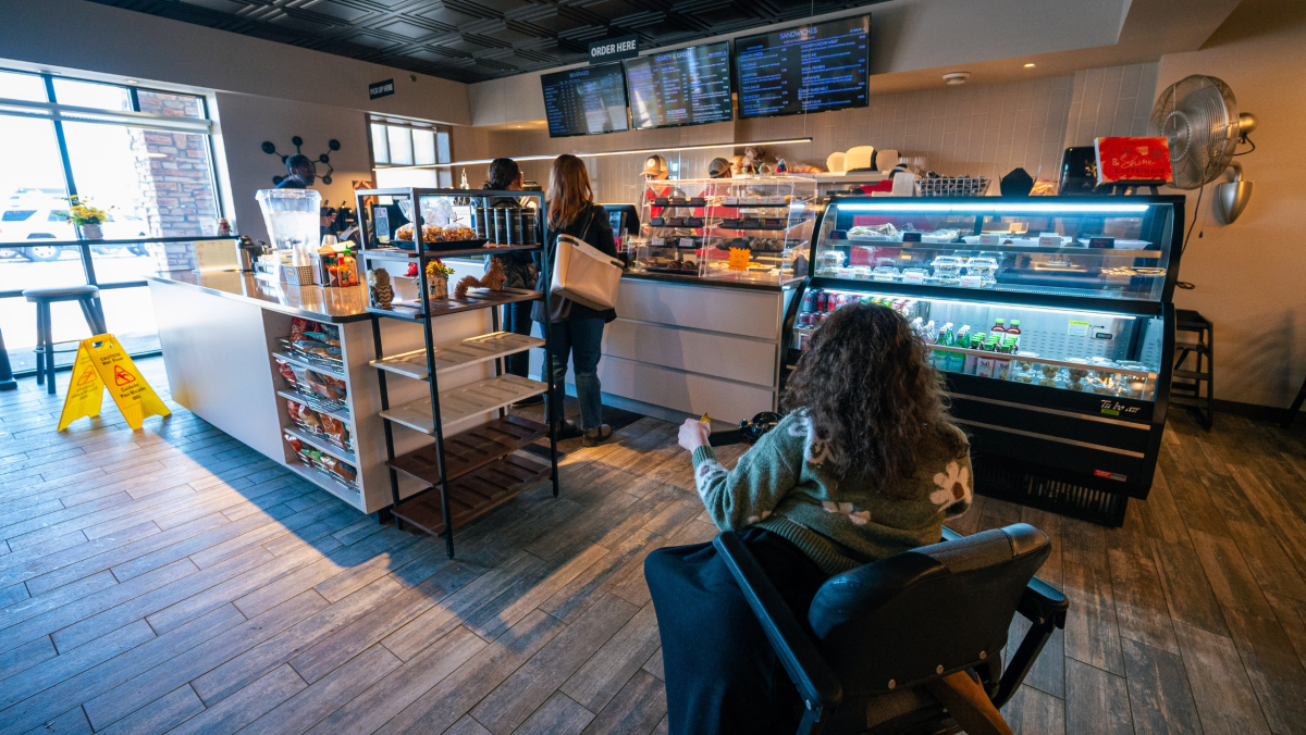 A cozy café interior with customers lined up at the counter. A person in a wheelchair waits, surrounded by displays of pastries and drinks, creating a warm, inviting atmosphere.