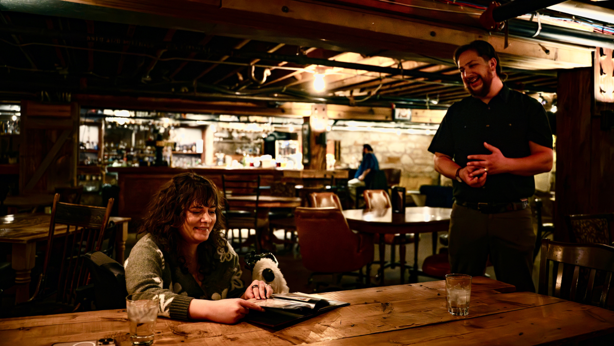 A cozy restaurant setting with dim lighting. A woman smiles at a menu on a wooden table, while a man stands nearby, engaged in conversation. Warm, friendly atmosphere.