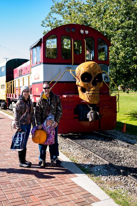 Family of three standing in front of the Pumpkin Train at the National Railroad museum smiling.