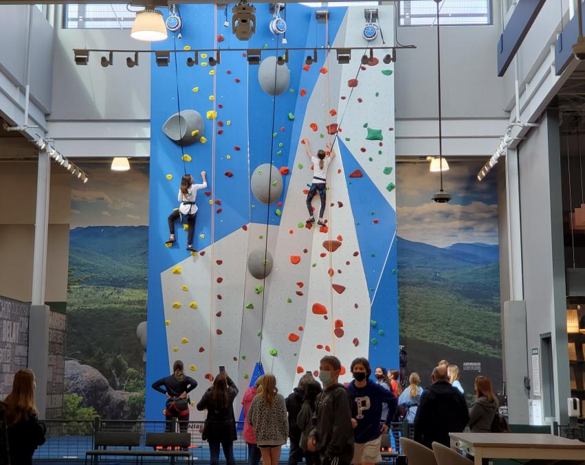 Two people climbing a blue and gray rock wall while a group of people watch at the bottom