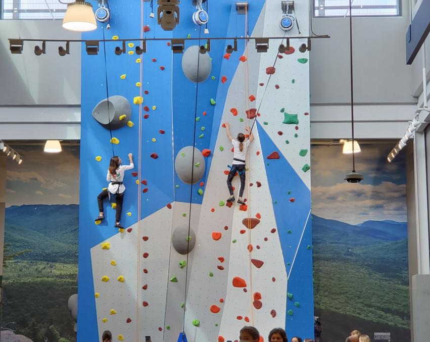 Two people climbing a blue and gray rock wall while a group of people watch at the bottom