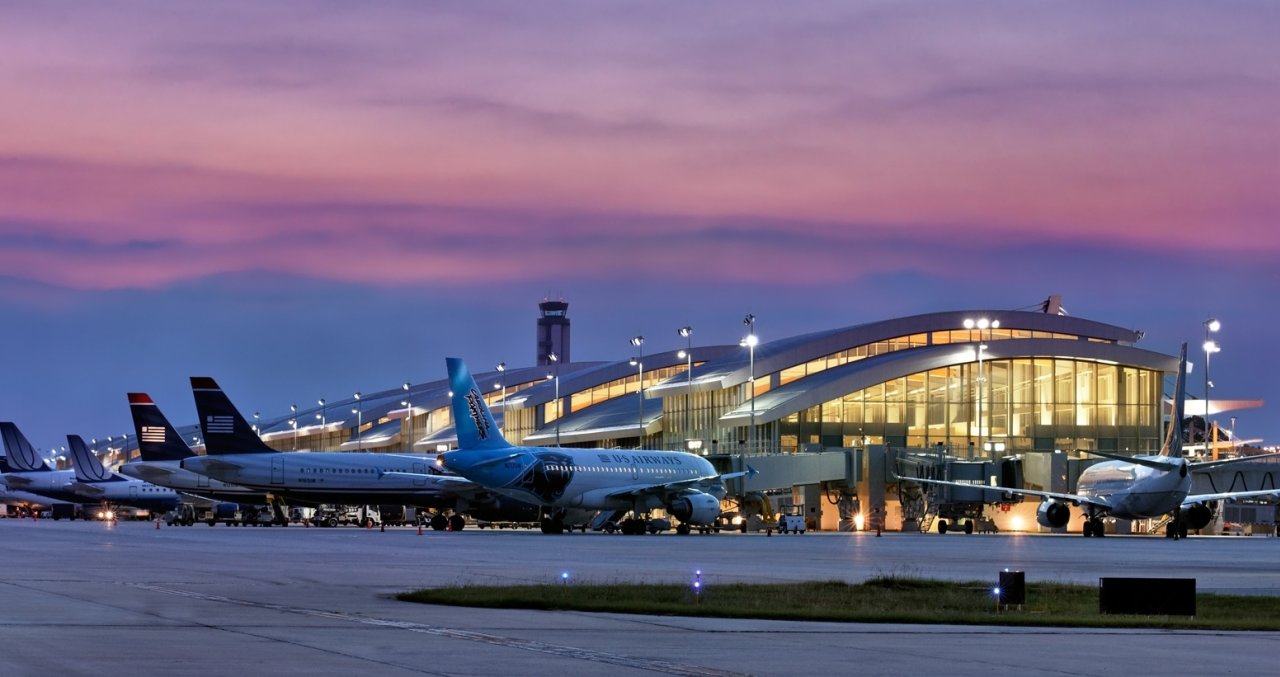 An airport scene at dusk, featuring several parked airplanes and a modern terminal illuminated against a colorful sky.