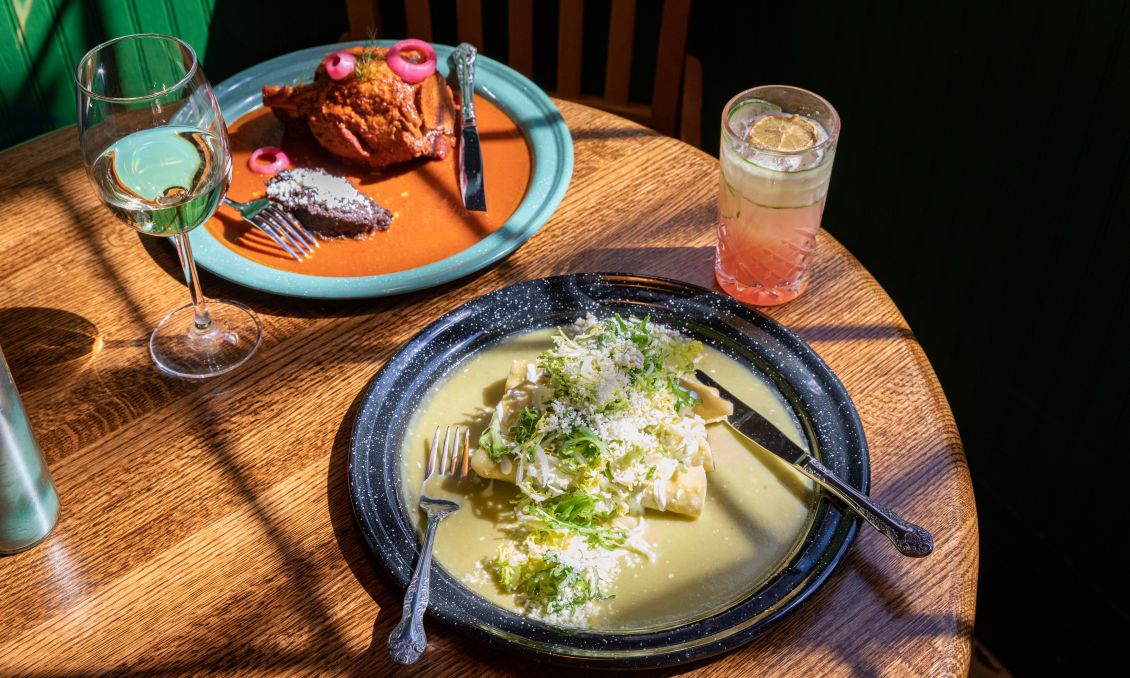 A 45-degree view of a restaurant table draped in sunlight with two dishes and two drinks; one plate has green chile chicken enchiladas and the other has Yucatan-style barbecued pork; drinks are a glass of white wine and a pinkish cocktail in a highball glass