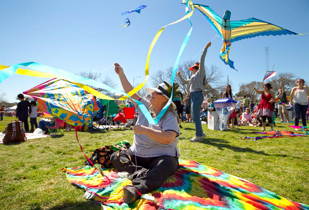 Woman sitting on rainbow-colored blanket in a grassy park, flying a kite.