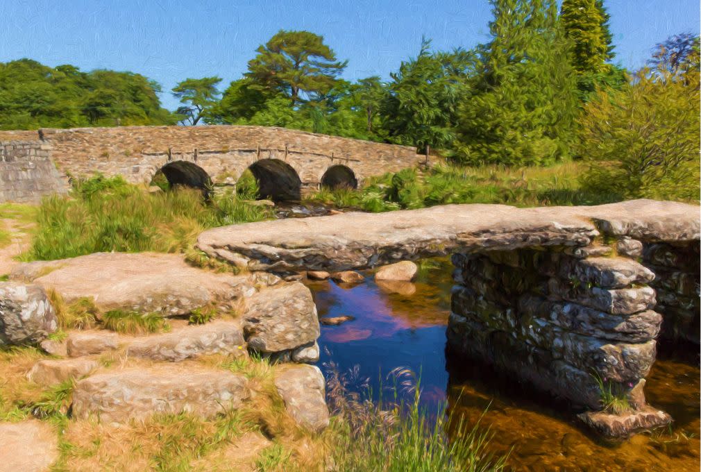 A picturesque stone bridge with three arches spans a gentle stream, surrounded by lush green trees and rocks under a clear blue sky. Calm and serene atmosphere.