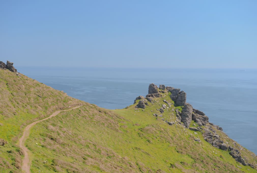 Coastal path on a grassy cliff leads to rugged rocks overlooking a calm, expansive blue sea under a clear sky, evoking a sense of tranquility