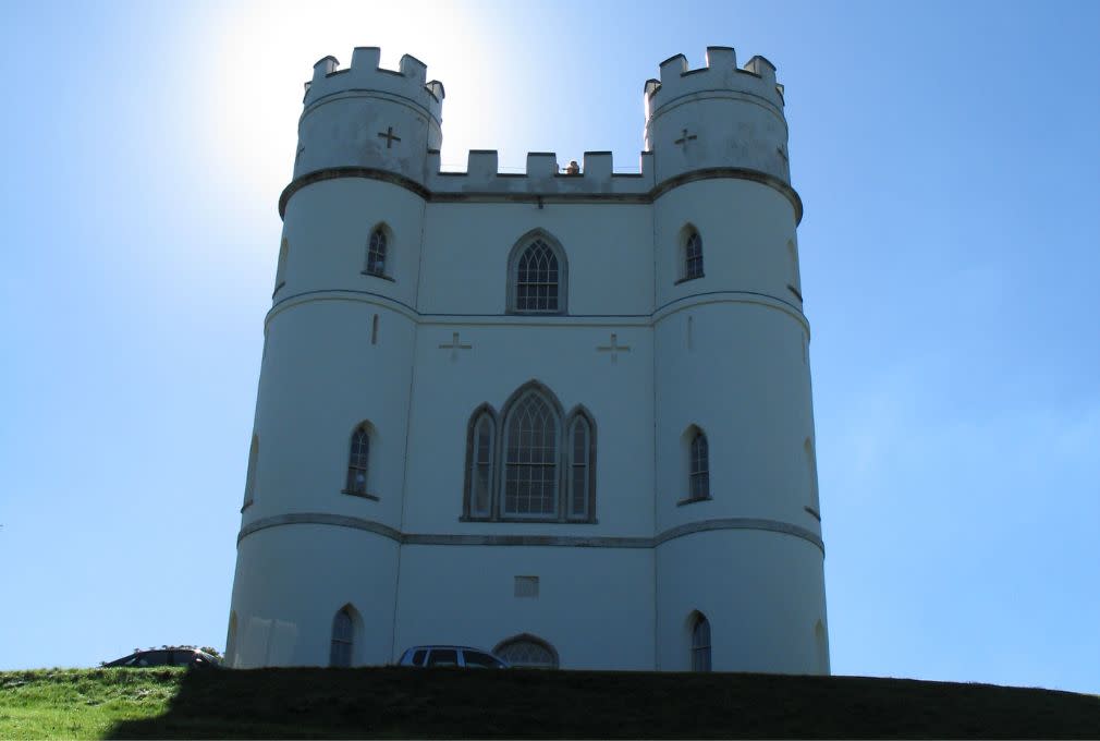 A white, castle-like tower with crenellations stands against a bright blue sky, the sun creating a halo effect. A car is parked on the grassy hill below.