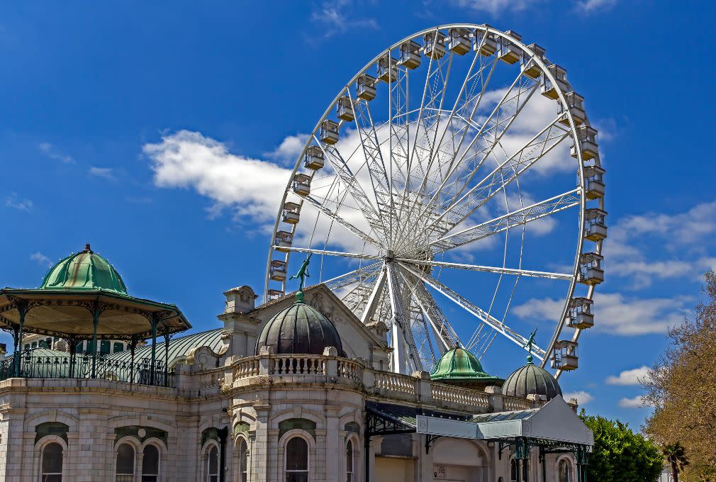 A large Ferris wheel rises behind an ornate historic building with domed green roofs under a bright blue sky with scattered clouds. A lively, whimsical atmosphere is conveyed.