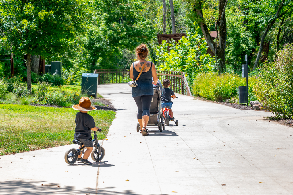 Sunny day on a paved trail with a mom pushing a stroller and kids riding bikes.