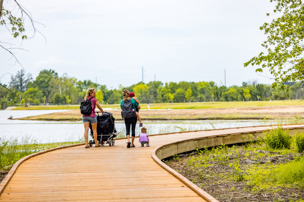 Two people with a stroller and small children walk on boardwalks over wetlands on a sunny day.