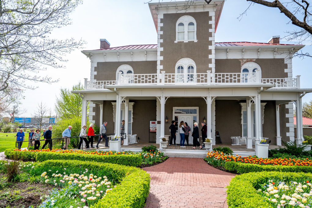 A stately Italianate home with a red brick path bordered by green shrubs and spring flowers.
