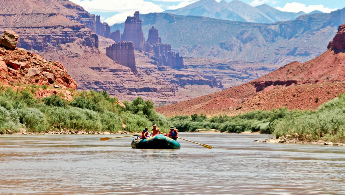 People in a teal raft surrounded by towering red rocks raft down the Colorado River near Moab.