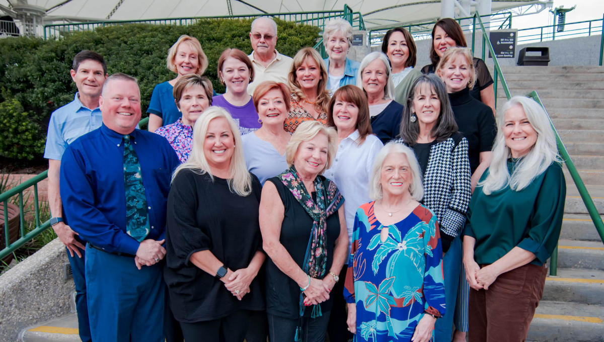 A group of 19 people (16 women and 3 men) smile at the camera. They are all dressed in business casual attire and standing on the steps leading up to The Pavilion concert seats.