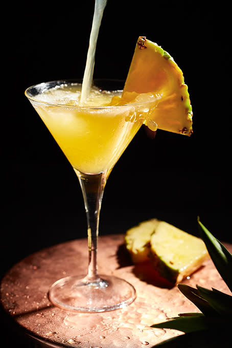 A martini glass with pineapple-like juice being poured into it and a slice of pineapple wedge on the edge of the glass as garnish.