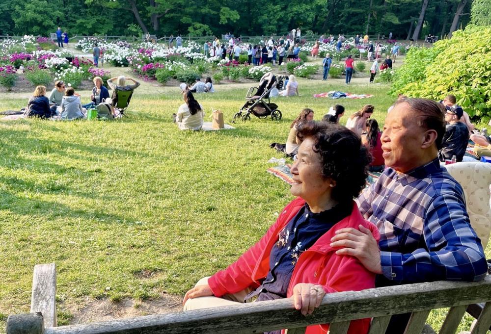Couple sits on wooden bench close to each other, looking to the left and smiling. They are at an outdoor garden with vibrant green grass and white and pink flowers in the background.