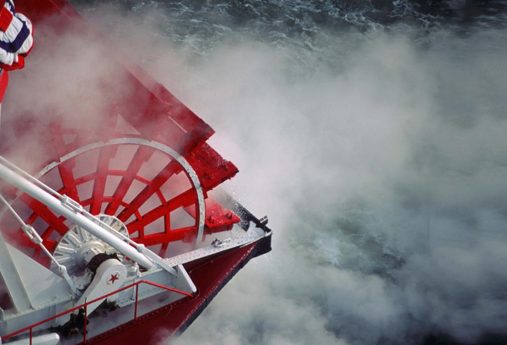A red paddlewheel on a red and white boat. Steam and mist churn up around the wheel.