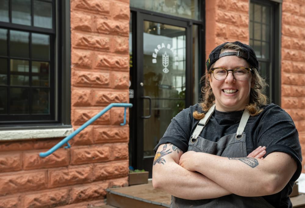 Woman wearing an apron and backwards baseball cap stands in front of business with her arms crossed and smiles at the camera.