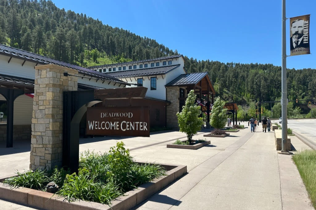 Deadwood Welcome Center on a sunny day, featuring a modern stone-and-wood building with a large “Deadwood Welcome Center” sign in front. A wide sidewalk lined with small trees leads toward the entrance, while forested hills rise behind the building under a clear blue sky. Several people are walking along the street, and a banner displaying a historical portrait hangs from a nearby light pole.