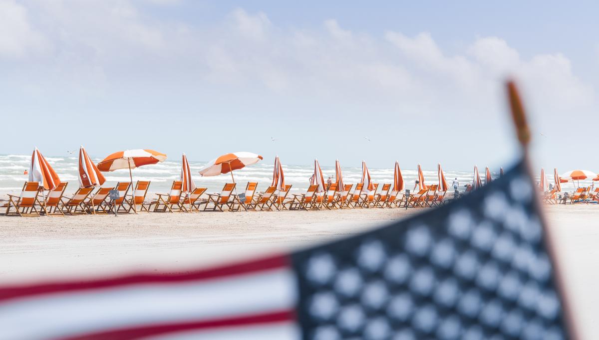 In focus is the background of the photo, which shows a row of orange and white beach chairs and umbrellas on the beach. An American flag is in the foreground, blurred.