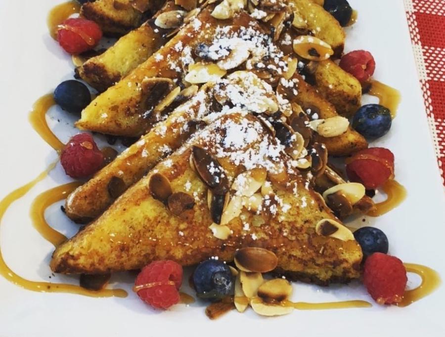 Four triangle-shaped slices of French toast layered on a plate topped with blue berries, raspberries, slivered almonds, powdered sugar and a drizzle of maple syrup