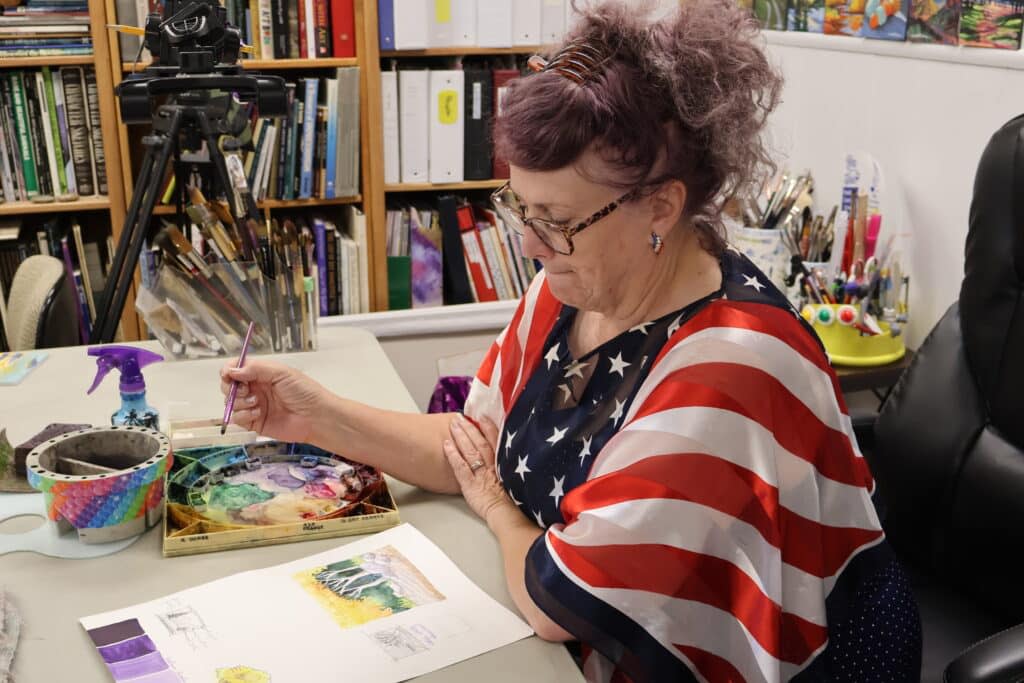 Colette Pitcher sits at a desk, looking down at an in-progress watercolor in shades of purple.