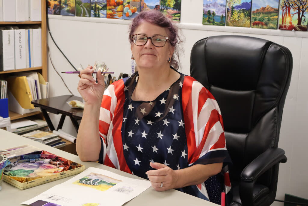 Colette Pitcher sits at a desk in her studio with a thin paintbrush in her hand.