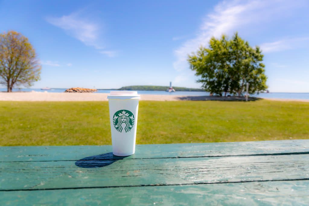 A cup of Starbucks coffee sits on a green picnic table at Mackinac Island's Windermere Point with water and lighthouse in background