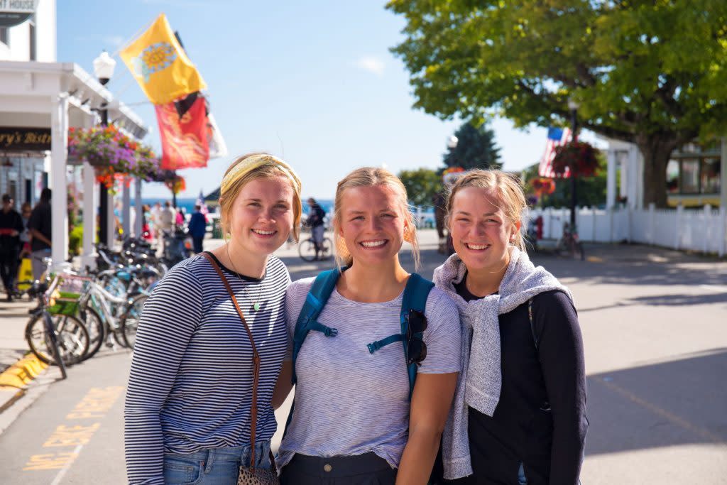 Three young women smile for the camera on Mackinac Island's Main Street on a sunny day.