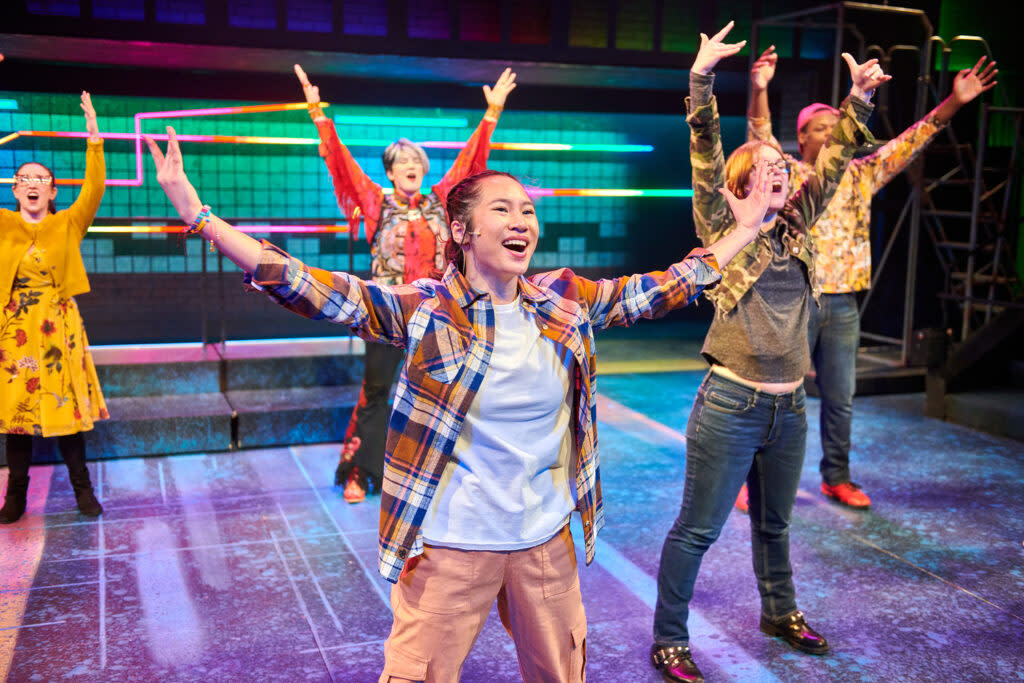 A group of young performers on a brightly lit stage throw their arms up mid-dance, smiling and full of energy, with colorful set pieces glowing behind them.