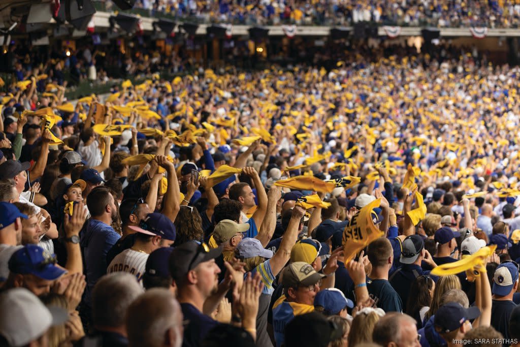 A packed stadium crowd cheers and waves yellow rally towels during a baseball game, filling the stands with motion and team colors.