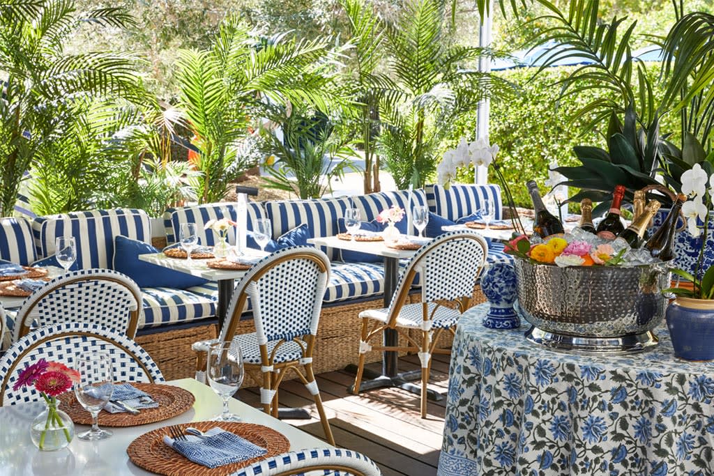 An outdoor dining area decorated with a blue and white color scheme, featuring striped couches, wicker chairs with blue patterned cushions, and a table set with plates and wine glasses. A metal ice bucket with champagne bottles is at the center, surrounded by green potted plants.