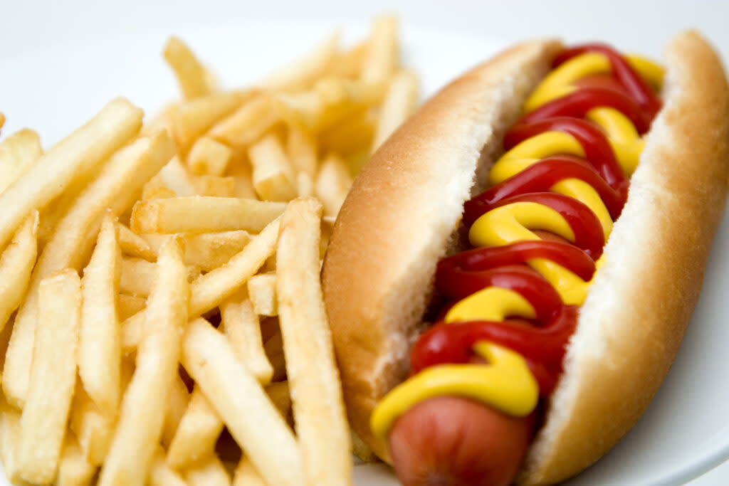 A hot dog with mustard and ketchup on a bun next to a pile of French fries on a white plate.